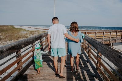 Family of four walking on a wooden boardwalk toward the beach, holding hands and looking at The Gulf.