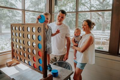 Family playing a large indoor Connect Four game together, with parents watching and smiling as a young child drops a game piece while holding a baby nearby