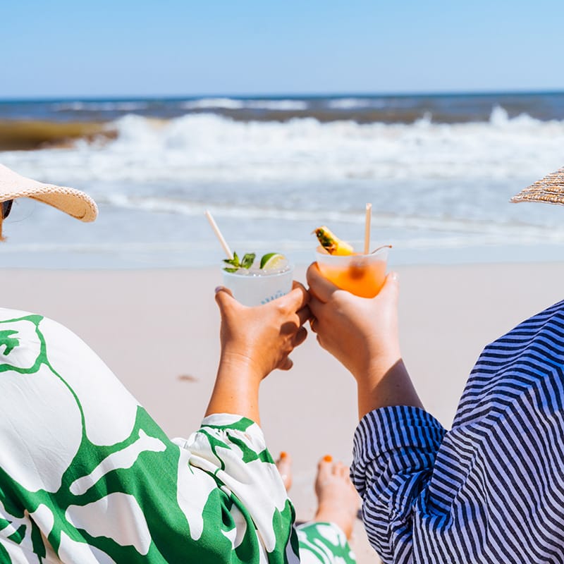 Two friends enjoying tropical drinks on a beach, with gentle waves in the background at a Gulf Coast resort