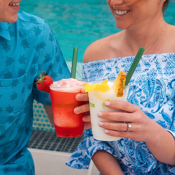 Couple relaxing poolside while holding tropical frozen drinks with fruit garnishes.