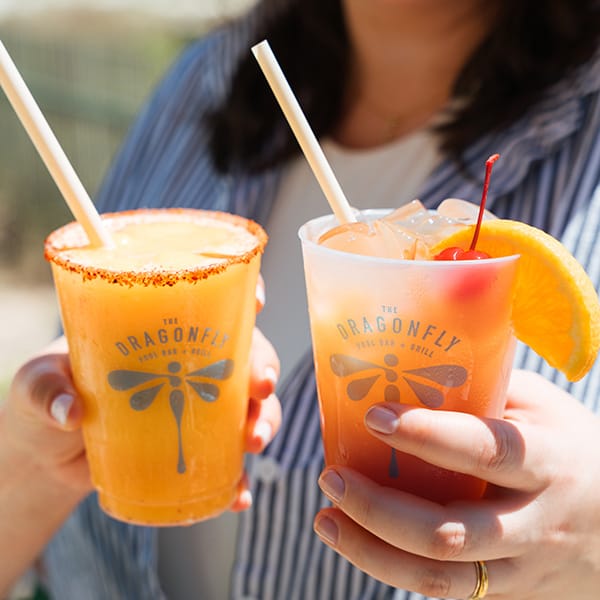Person holding two colorful frozen cocktails garnished with fruit at an outdoor bar.
