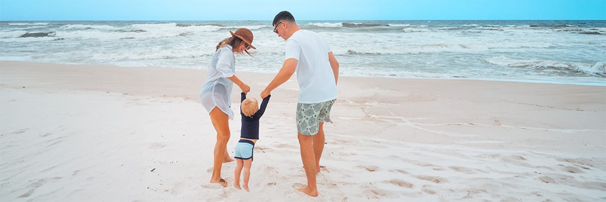 Parents swinging a young child by the hands while playing together on a sandy beach near the ocean.