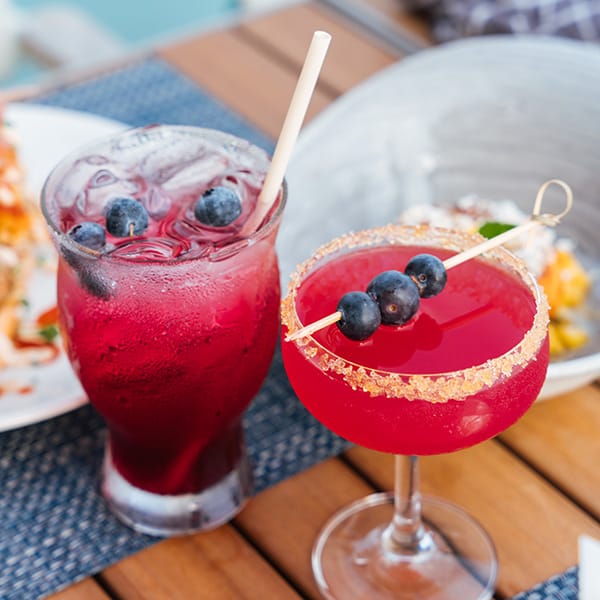 Two vibrant berry cocktails served on a wooden restaurant table with fresh blueberries as garnish.