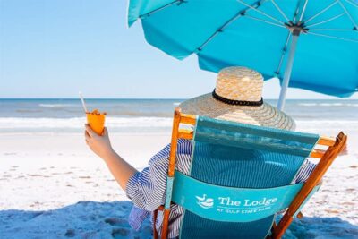 Person relaxing in a beach chair under a turquoise umbrella while holding a refreshing drink by the ocean.